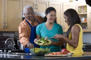 Familia preparando una ensalada juntos. La madre está sirviendo a su hija y el padre está comiendo una verdura del bol.