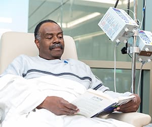 Man sitting in chair, reading book while getting I.V. chemo treatment.